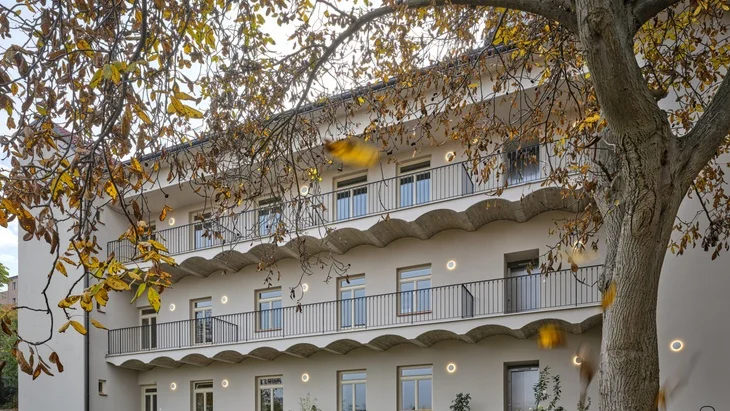 Bílý dům s balkony a okny částečně zakrytý listím stromu.