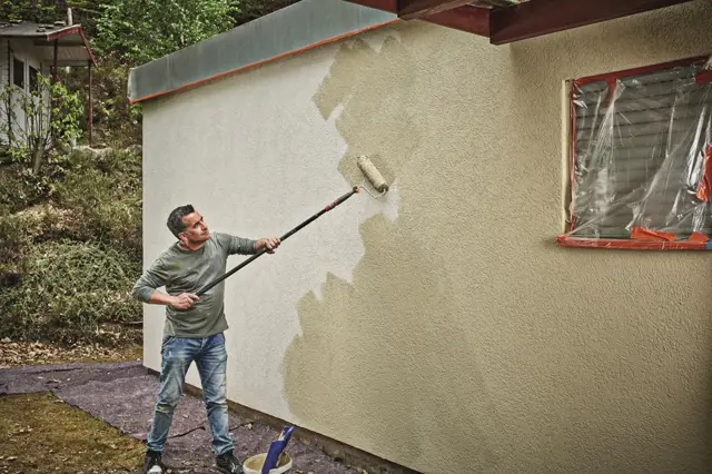 Muž maluje fasádu hnědou barvou u okna.