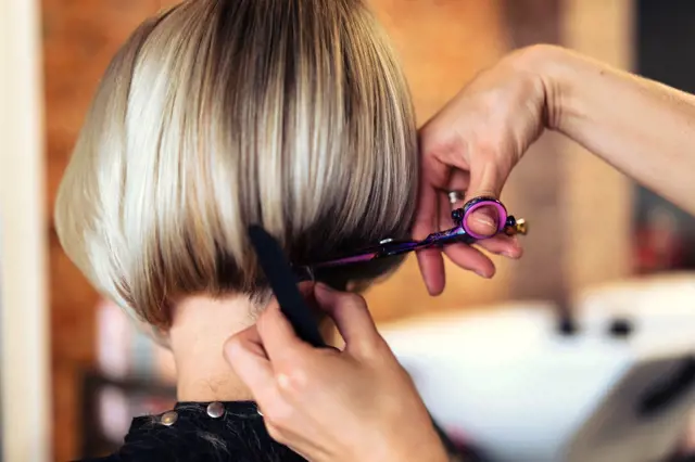 Stylista stříhá blond účes podkovu zezadu.
