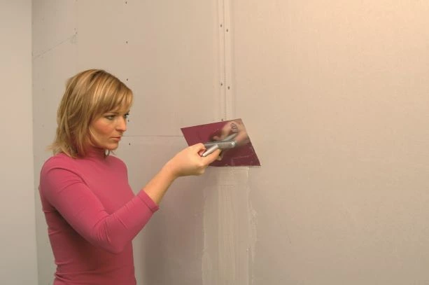 Žena nanáší tmel na sádrokartonovou stěnu špachtlí.