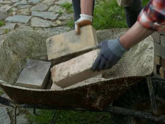 Ruce s rukavicemi nakládají cihly do vozíku.