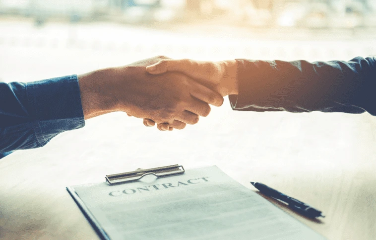 Ruce se stiskají nad dokumentem s nápisem CONTRACT.