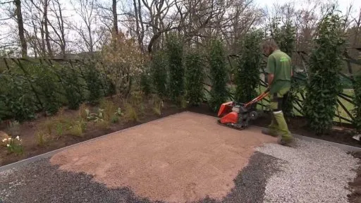 Muž tluče štěrkovou plochu vibračním válcem v zahradě.